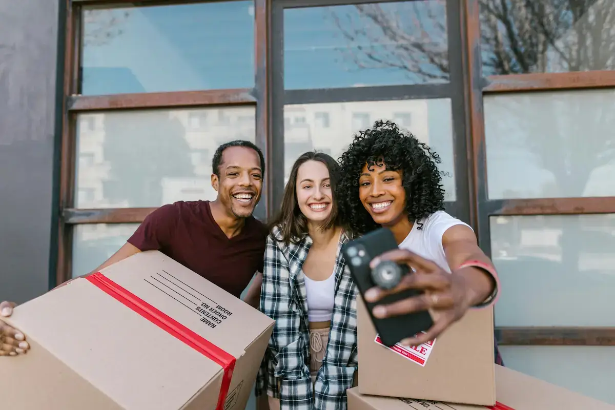 Three college students smiling and carrying moving boxes into a university dorm room during move-in day.