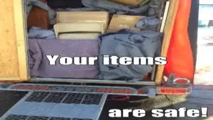 A perfectly packed U-Box portable storage container sits securely on a tree-lined residential driveway of a triple-decker apartment building in Massachusetts. The container door is open, revealing a "professional load." A solid wall of uniform moving boxes forms a flat foundation across the width of the container. Above them, larger items like a mattress (protected in a bag), a disassembled wooden table, and wrapped furniture are precision-fitted. Moving blankets and smaller soft items are strategically wedged to "chock" the load, filling every void and ensuring zero gaps. The loading is neat, balanced, and contained completely within the structure, highlighting professional packing technique in a challenging urban setting. In the background, typical densely packed wooden residential homes and a green street sign with local text emphasize the tight urban environment where efficient packing is critical.
